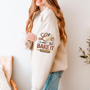 Pode incluir: Camisola bege com o texto "Life is what you bake it" em letras castanhas e amarelas, juntamente com um gr&aacute;fico de rolo de massa. A camisola &eacute; de cor neutra e tem mangas compridas.