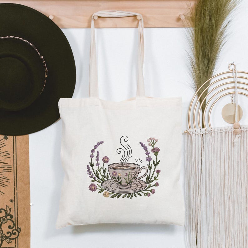 May include: A cream tote bag with an embroidered teacup design. The teacup is surrounded by purple lavender and other flowers. The bag has two cream handles and is hanging on a wooden rack.