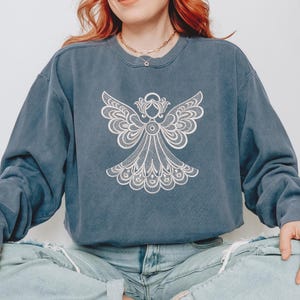 May include: A blue-grey sweatshirt featuring a detailed white angel design on the front. The angel has outstretched wings and intricate lace-like detailing. The sweatshirt has a crew neck and long sleeves, with a slightly faded appearance.
