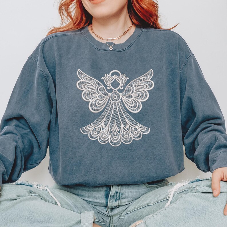 May include: A blue-grey sweatshirt featuring a detailed white angel design on the front. The angel has outstretched wings and intricate lace-like detailing. The sweatshirt has a crew neck and long sleeves, with a slightly faded appearance.
