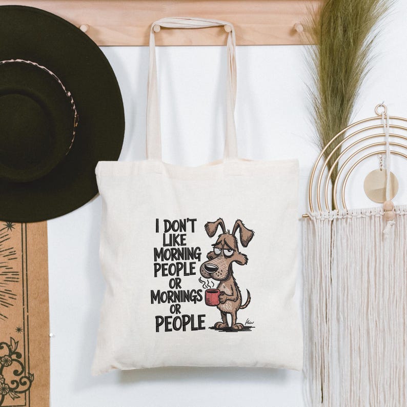 May include: Cream-colored tote bag with the text "I DON'T LIKE MORNING PEOPLE OR MORNINGS OR PEOPLE." A cartoon dog with a grumpy expression holds a red mug. The bag has two cream-colored handles.