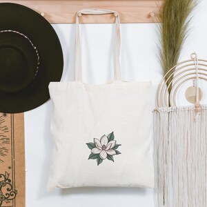 Pode incluir: Saco tote creme com um design de flor de magn&oacute;lia. A flor &eacute; rosa e branca com folhas verdes. O saco est&aacute; pendurado em um cabide de madeira, com um chap&eacute;u preto e outros itens decorativos no fundo.