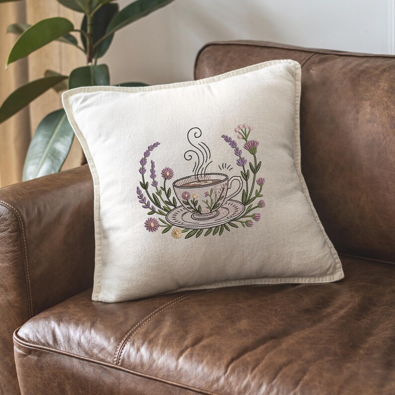 May include: Cream-coloured square cushion featuring an embroidered design of a steaming teacup and saucer, encircled by a floral wreath of lavender and other flowers. The cushion is on a brown leather sofa.