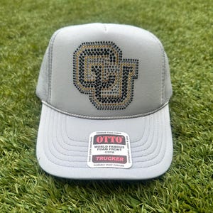 May include: A gray trucker hat with a black and gold rhinestone OU logo. The hat has a mesh back and a snap closure. The hat is labeled "OTTO World Famous Foam Front Trucker".