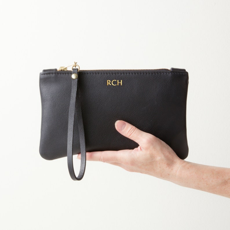 Hand holding monogrammed leather clutch with wrist strap.
