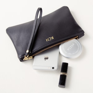 Monogrammed leather clutch with wrist strap angled view with cell phone and makeup partially inside clutch.