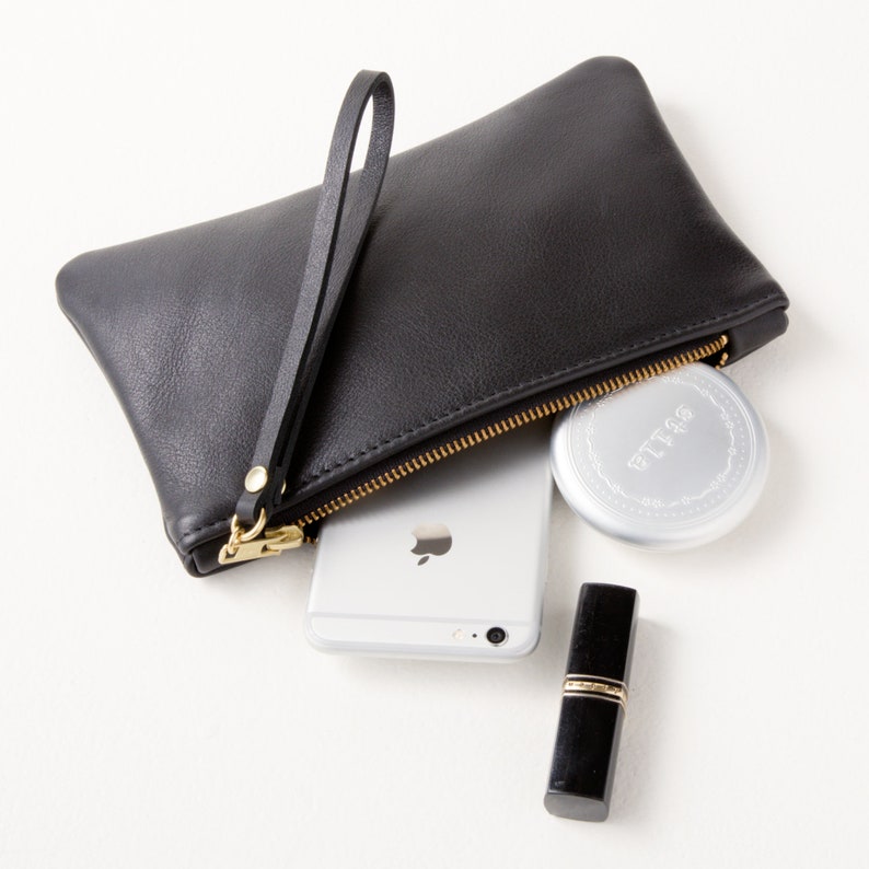 Black leather zipper clutch with wrist strap angled view with cell phone and makeup partially inside clutch.