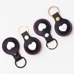 May include: Four black leather keychains with heart-shaped cutouts and metal key rings. Each keychain has a different color of stitching around the heart: red, pink, and purple. The keychains have gold, silver, and black hardware.