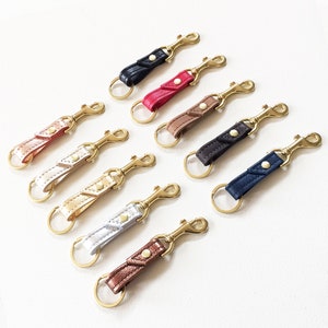May include: Ten leather keychains with gold metal clasps and rings. The keychains are in various colors, including black, red, brown, beige, gold, and silver.