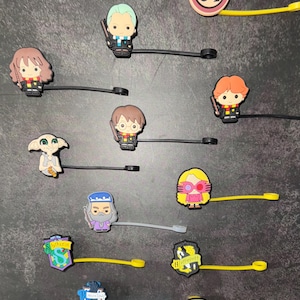 Puede incluir: Una colección de coloridos amuletos de goma con personajes y símbolos de la serie Harry Potter. Los amuletos incluyen personajes como Harry, Hermione y Dobby, así como los escudos de las casas de Hogwarts y el letrero de la plataforma 9 3/4.