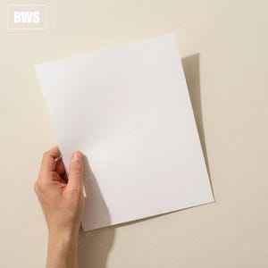 May include: A hand holding a blank white sheet of paper, with the letters "BWS" in the upper left corner. The paper is positioned at an angle against a neutral background, creating a shadow.