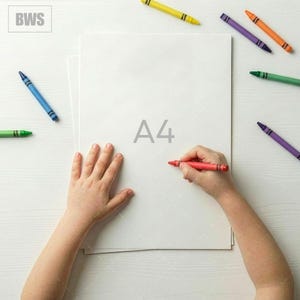 May include: A top-down view of a person's hands drawing on a white A4 paper with a red crayon. Several colorful crayons, including yellow, blue, green, orange, and purple, are scattered around the paper on a white wooden surface.