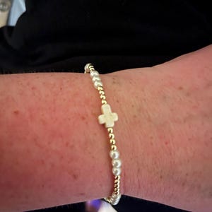 Op de afbeelding: Een armband met een witte kruisbedel, gouden kralen en witte parels.