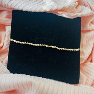 May include: A gold beaded bracelet on a black background. The bracelet is made of small, round beads.