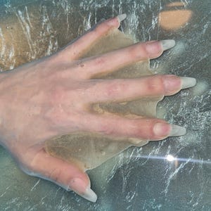 Könnte beinhalten: Eine durchscheinende, beige, flexible Silikonform oder Abdeckung in Handform mit glatter Textur. Das Objekt ist so geformt, dass es über Handfläche und Finger passt, und besteht aus einem dünnen, biegsamen Material, das sich der Handform anpasst.