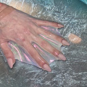 May include: A clear, iridescent hand accessory with extended, pointed finger covers. The accessory has a pearlescent sheen with pink and green hues. The nails are painted a pale pink color. The accessory is designed to fit over the hand.