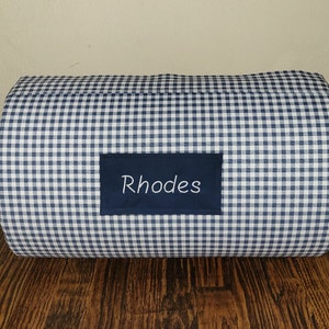 May include: A rectangular, navy blue and white gingham patterned pillow. A navy blue rectangular patch is sewn onto the pillow and features the embroidered name "Rhodes" in white script.