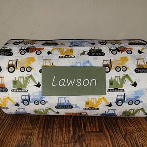 May include: A rectangular pillow with a white background featuring colorful illustrations of construction vehicles. A green rectangular patch with the name "Lawson" in white script is centered on the pillow. The pillow is approximately 12 inches long.
