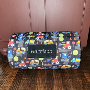 May include: A rectangular bolster pillow with a black background and a colorful pattern of trucks, trees, and road signs. The name "Harrison" is embroidered on a black patch.