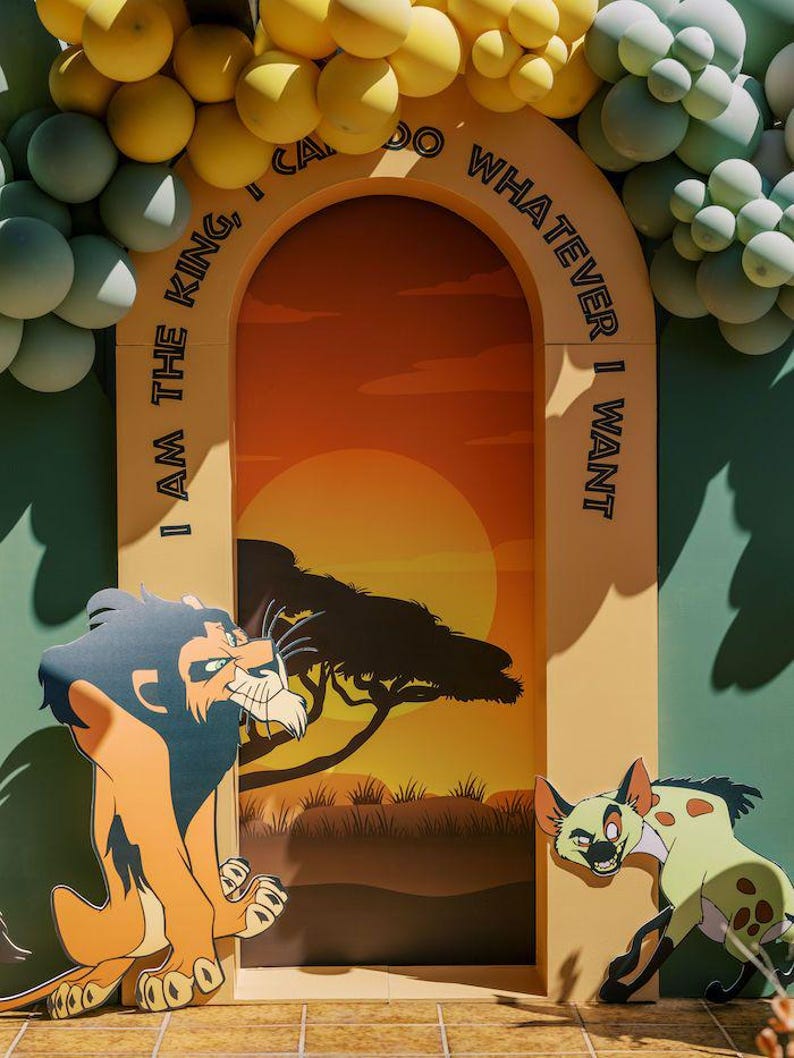 May include: A cardboard archway decorated with yellow and green balloons. The archway is decorated with the text "I am the king, I can do whatever I want".  The archway is set up in front of a backdrop depicting a sunset over a savanna with a lion and a hyena.