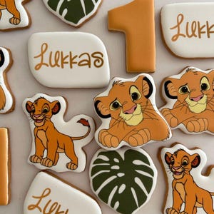 May include: A set of decorated sugar cookies featuring a lion character, a jungle leaf, and the name "Lukkas" written in gold lettering. The cookies are decorated with royal icing and edible images.