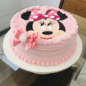 Pode incluir: Um bolo rosa e branco decorado com uma imagem da Minnie Mouse. O bolo tem um la&ccedil;o rosa e detalhes de p&eacute;rolas brancas.