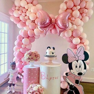 Pode incluir: Um arco de bal&otilde;es rosa e branco em forma de orelhas do Mickey Mouse com um grande la&ccedil;o rosa no centro. Dois suportes para bolos brancos com bolinhas rosa, um com um bolo rosa e branco decorado com uma cabe&ccedil;a do Mickey Mouse e o n&uacute;mero 2. Um suporte para bolos rosa e branco com bolinhas com o nome "Blythe" escrito nele. Um grande recorte de papel&atilde;o da Minnie Mouse com um vestido rosa com bolinhas brancas.