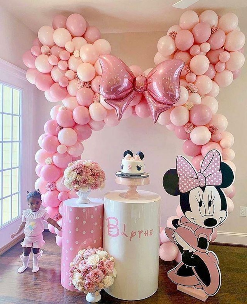 Pode incluir: Um arco de bal&otilde;es rosa e branco em forma de orelhas do Mickey Mouse com um grande la&ccedil;o rosa no centro. Dois suportes para bolos brancos com bolinhas rosa, um com um bolo rosa e branco decorado com uma cabe&ccedil;a do Mickey Mouse e o n&uacute;mero 2. Um suporte para bolos rosa e branco com bolinhas com o nome "Blythe" escrito nele. Um grande recorte de papel&atilde;o da Minnie Mouse com um vestido rosa com bolinhas brancas.