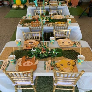 May include: A white table with a gold glitter table runner and green palm leaves. The table is set with gold chairs, paper cups, and plates with cartoon animal faces. The plates feature Simba, Pumbaa, and Timon from the Lion King.