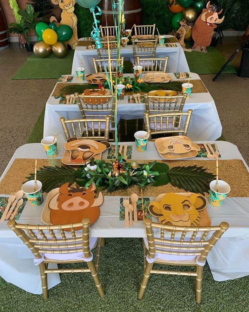 May include: A white table with a gold glitter table runner and green palm leaves. The table is set with gold chairs, paper cups, and plates with cartoon animal faces. The plates feature Simba, Pumbaa, and Timon from the Lion King.