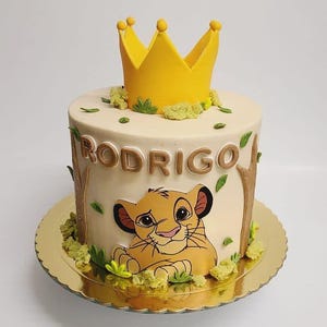May include: A white cake decorated with a yellow crown, green leaves, and a cartoon image of Simba from The Lion King. The cake is on a gold cake stand and says "RODRIGO" in brown letters.