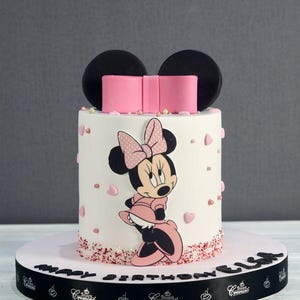 Pode incluir: Um bolo branco decorado com um la&ccedil;o rosa, orelhas pretas e uma imagem de desenho animado do Mickey Mouse. O bolo &eacute; cercado por confeitos rosa e brancos e cora&ccedil;&otilde;es. O bolo est&aacute; em um tabuleiro de bolo branco com uma fita preta que diz "Feliz anivers&aacute;rio" e o nome "Lila".