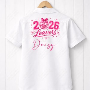 Op de afbeelding: Wit shirt met korte mouwen en een roze afbeelding op de rug. De afbeelding bevat de nummers "2026", het woord "Leavers", een discobal en de naam "Daisy". Het shirt hangt aan een houten hanger.