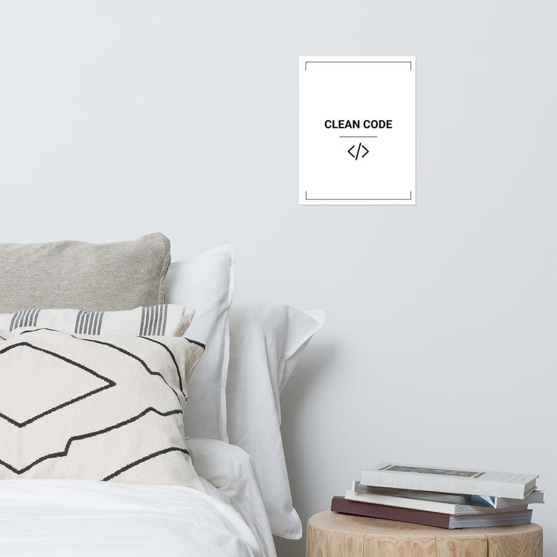 May include: A rectangular white wall poster featuring a thin black border, the text CLEAN CODE in black capital letters, and a black code bracket symbol centered below the text. The poster is displayed on a plain white wall above a bed.