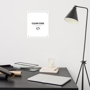 May include: A white rectangular wall poster featuring a thin black border, the text CLEAN CODE in black capital letters, and a black code bracket symbol centered below the text. The poster is displayed on a plain white wall above a desk.