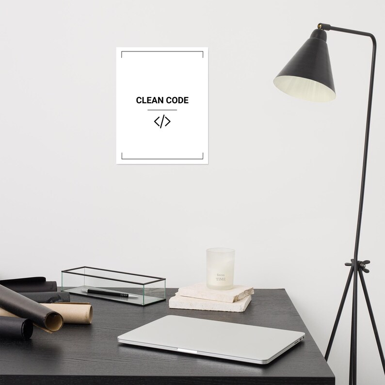 May include: A white rectangular wall poster featuring a thin black border, the text CLEAN CODE in black capital letters, and a black code bracket symbol centered below the text. The poster is displayed on a plain white wall above a desk.