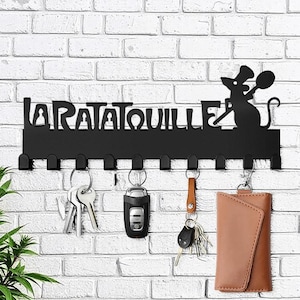 May include: Black metal key holder with the text "LA RATATOUILLE" and a silhouette of a chef rat. The key holder has several hooks with keys and a brown leather key case hanging from them. The key holder is mounted on a white brick wall.