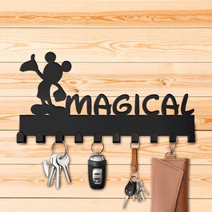 Könnte beinhalten: Schwarzer Schlüsselhalter aus Metall mit einer Silhouette von Mickey Mouse und dem Wort "MAGICAL". Der Halter hat 10 Haken und ist an einer Holzwand montiert.