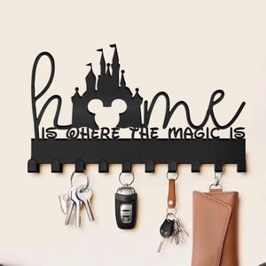 Könnte beinhalten: Schwarzer Schlüsselhalter aus Metall mit einer Silhouette eines Schlosses und Mickey-Maus-Ohren. Der Text "home is where the magic is" ist auf dem Halter geschrieben. Der Halter hat 10 Haken zum Aufhängen von Schlüsseln.