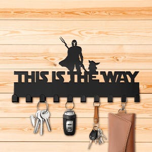 Könnte beinhalten: Schwarzer Schlüsselhalter aus Metall mit einer Silhouette eines Mandalorianers und Grogu, mit dem Text "This is the Way". Der Schlüsselhalter hat acht Haken und ist mit Schlüsseln und einer Brieftasche bestückt.
