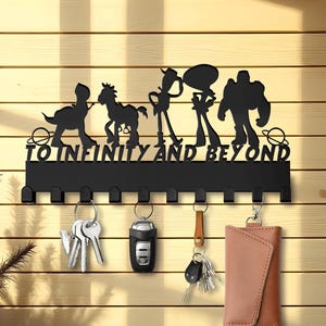 May include: Black metal key holder featuring a silhouette of Toy Story characters and the text "To Infinity and Beyond". The key holder has eight hooks and is mounted on a wooden wall.