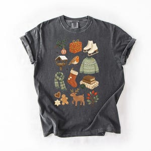 A gray t-shirt with a collage of various Christmas-themed items and decorations, including a pair of skis, a gift box, a Santa hat, a reindeer, a gingerbread man, and other festive symbols.