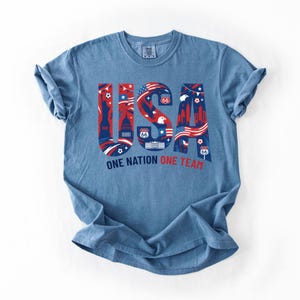 May include: A blue short-sleeve t-shirt featuring a large graphic of the letters USA in red, white, and blue. The letters contain illustrations of the Statue of Liberty, the White House, eagles, and Route 66 signs. Below is the text ONE NATION ONE TEAM.