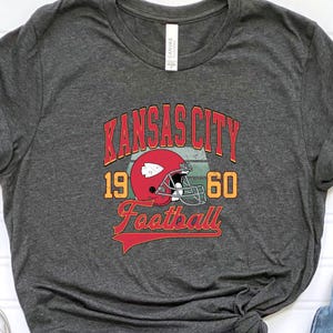 K&ouml;nnte beinhalten: Ein dunkelgraues T-Shirt mit einer Grafik im Vintage-Stil eines Footballhelms und dem Text "Kansas City 19 60 Football".