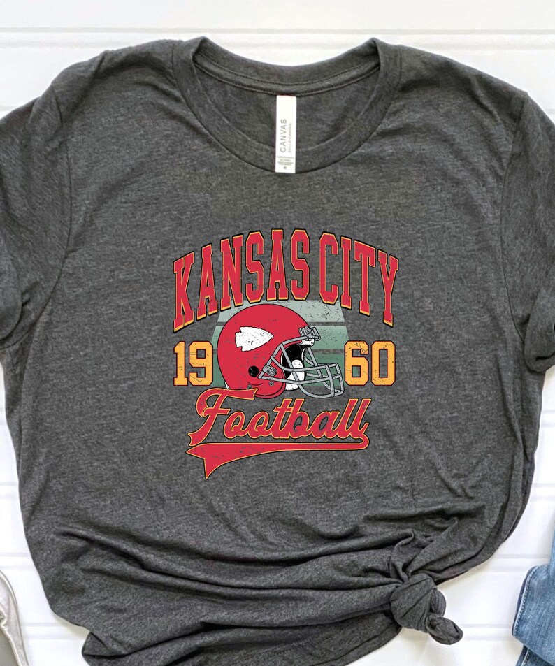K&ouml;nnte beinhalten: Ein dunkelgraues T-Shirt mit einer Grafik im Vintage-Stil eines Footballhelms und dem Text "Kansas City 19 60 Football".