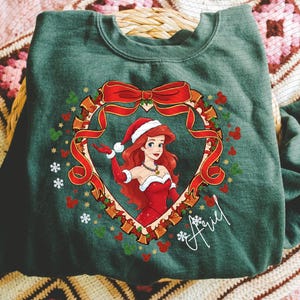 A green t-shirt with a Christmas-themed design featuring a cartoon character wearing a Santa hat and red dress, surrounded by a wreath and snowflakes.