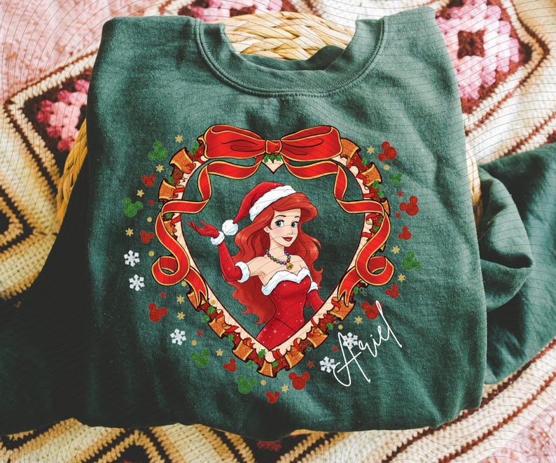 A green t-shirt with a Christmas-themed design featuring a cartoon character wearing a Santa hat and red dress, surrounded by a wreath and snowflakes.