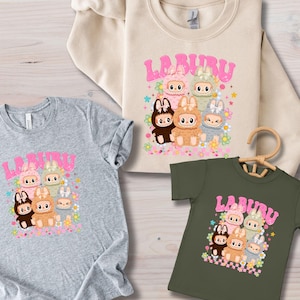 May include: Three shirts with a cartoon graphic of bunny-like characters and the word "LABUBU" in pink bubble letters. The shirts are gray, beige, and olive green. The graphic includes flowers and stars.