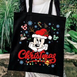 Puede incluir: Bolso tote negro con un dise&ntilde;o de Minnie Mouse con tem&aacute;tica navide&ntilde;a. Minnie lleva un gorro de Pap&aacute; Noel con cuernos, rodeada de copos de nieve, bastones de caramelo y luces navide&ntilde;as. La palabra "Christmas" est&aacute; escrita en rojo.