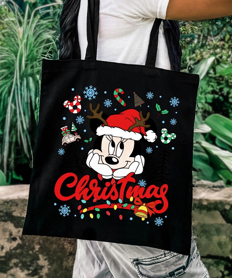 Puede incluir: Bolso tote negro con un dise&ntilde;o de Minnie Mouse con tem&aacute;tica navide&ntilde;a. Minnie lleva un gorro de Pap&aacute; Noel con cuernos, rodeada de copos de nieve, bastones de caramelo y luces navide&ntilde;as. La palabra "Christmas" est&aacute; escrita en rojo.
